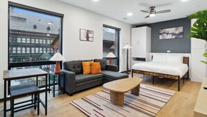 A modern living room with a sofa, coffee table, and a bed. There is a large window with a view of buildings outside. - CozySuites at Block20 (Indianapolis)