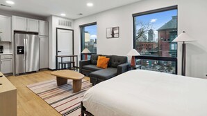 A modern apartment with a kitchenette, a sofa, a bed, and large windows offering a city view. - CozySuites at Block20 (Indianapolis)