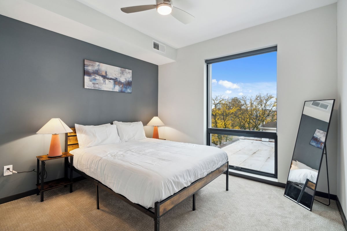 A modern bedroom with a large window, a bed, bedside lamps, and a ceiling fan.
