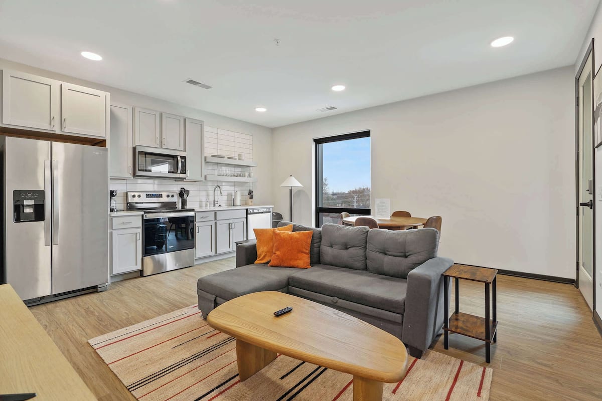 A modern apartment with a kitchen, a gray sectional sofa, a wooden coffee table, and a view of the city through a large window.