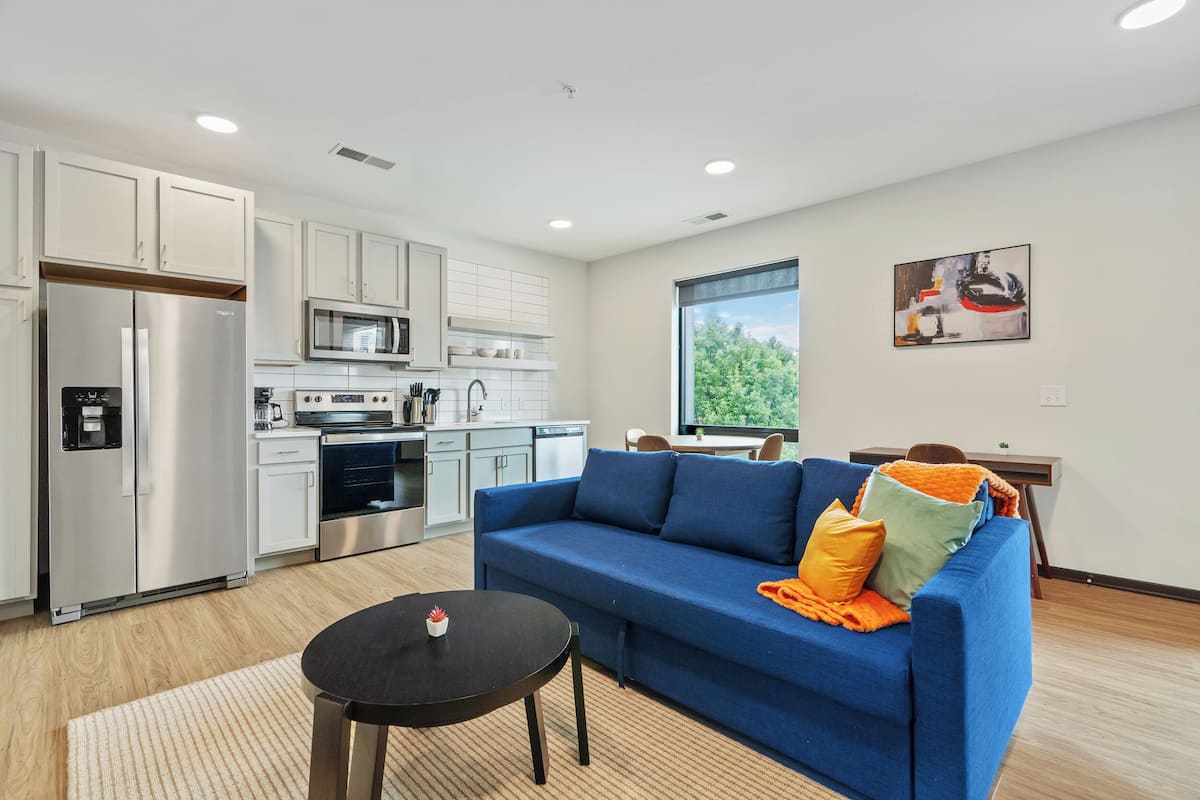 A modern kitchen with stainless steel appliances, a blue sofa with orange cushions, and a dining area with a table and chairs.