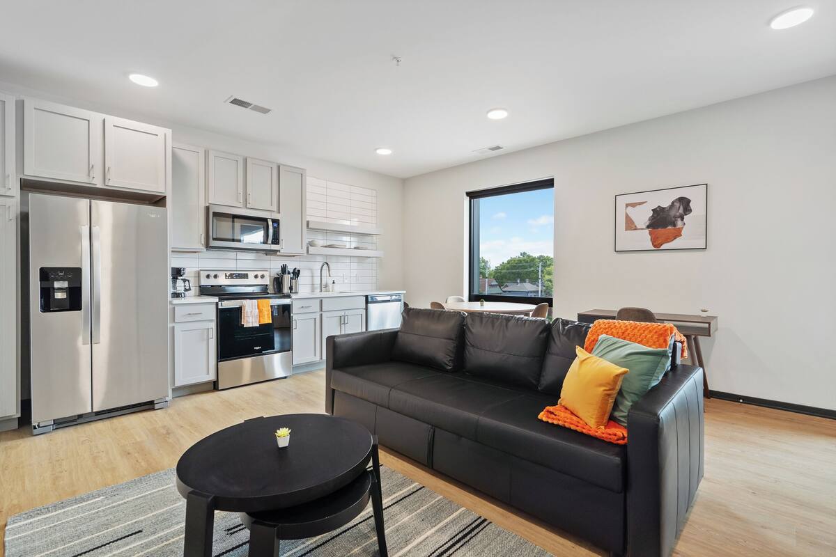 A modern kitchen with stainless steel appliances, a black leather sofa, and a dining table with a rug.