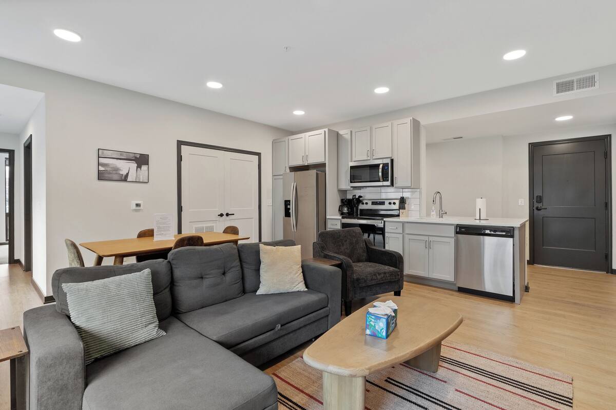 A modern living room with a sofa, armchair, and coffee table, a dining area with a table and chairs, and a kitchen with appliances and cabinets.
