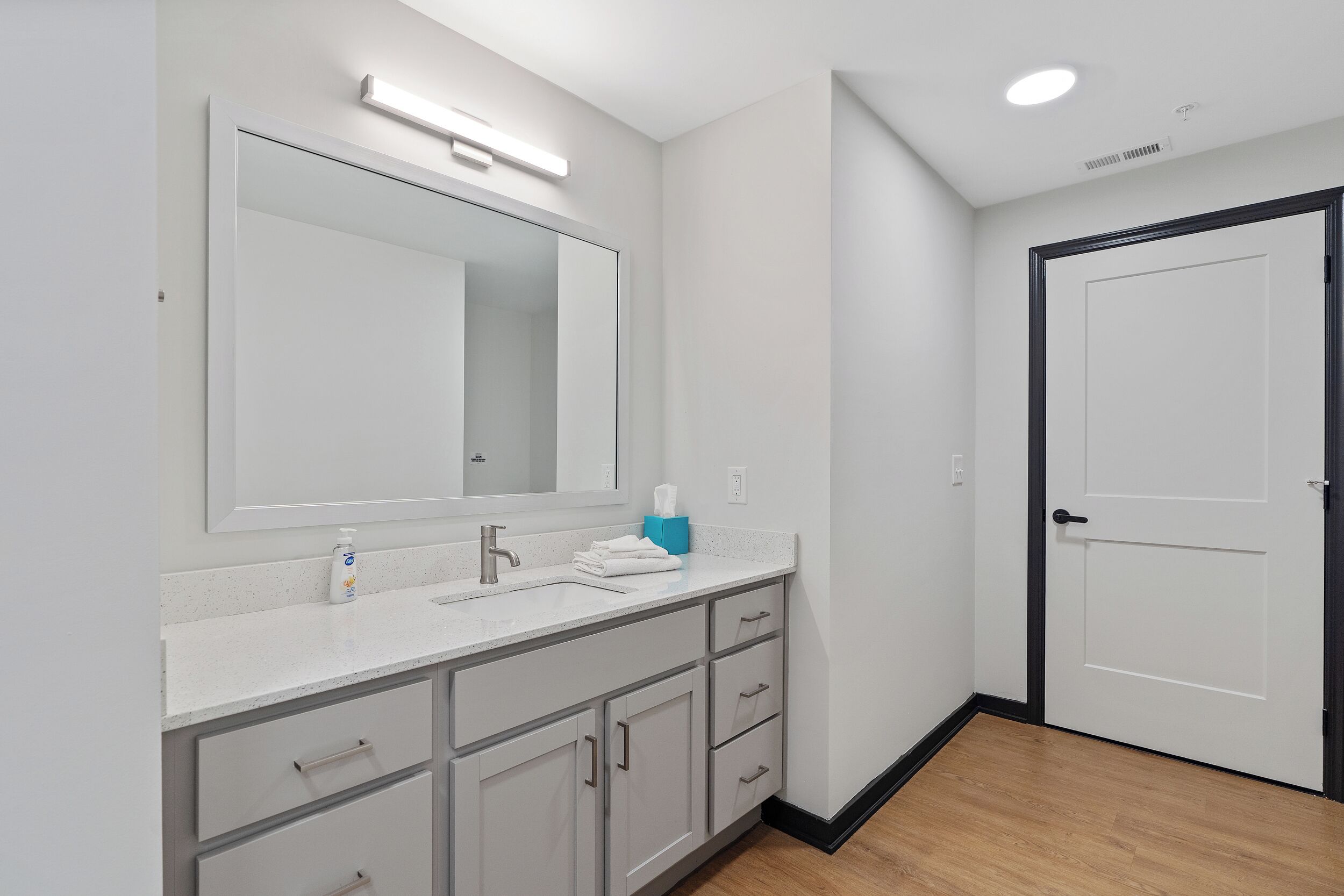 Un baño moderno con un espejo grande, encimera blanca y puerta con marco negro.