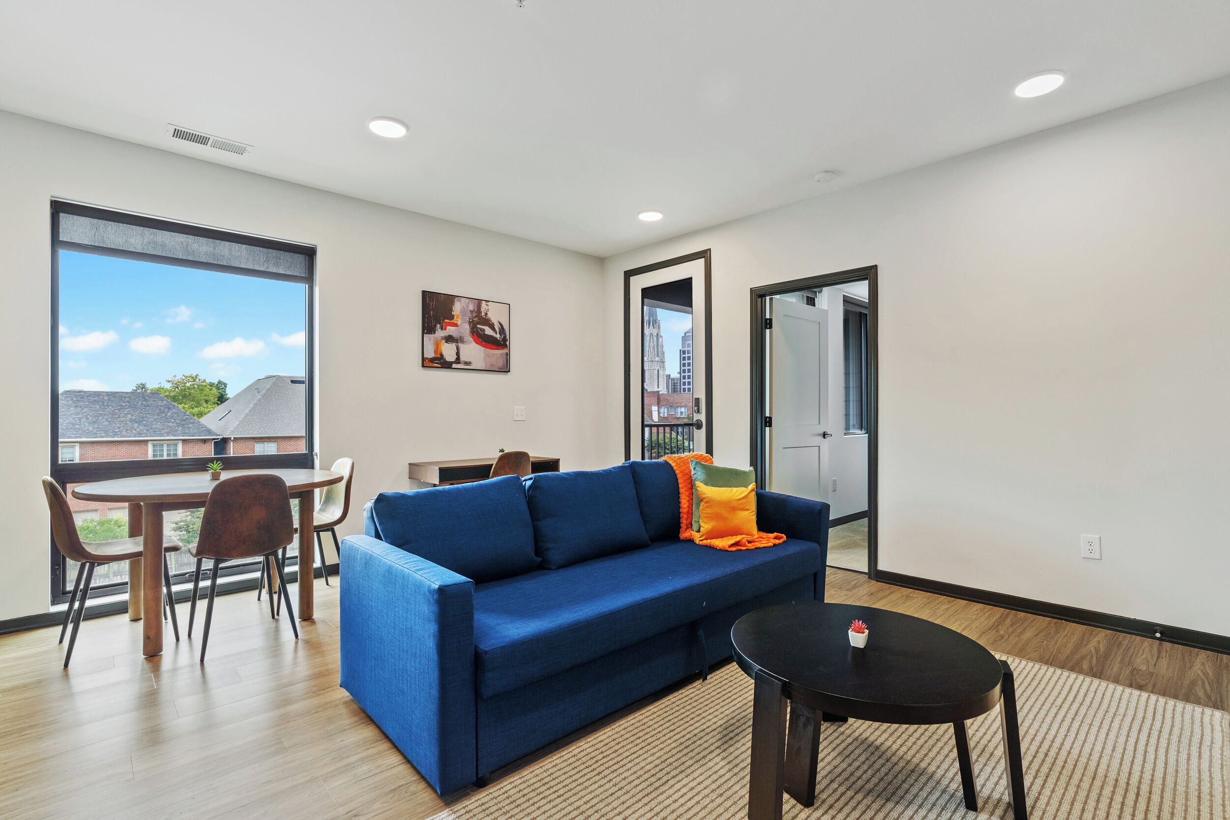 Een moderne woonkamer met een blauwe bank, een eettafel met stoelen, een kleine zwarte tafel en een groot raam met uitzicht op gebouwen.