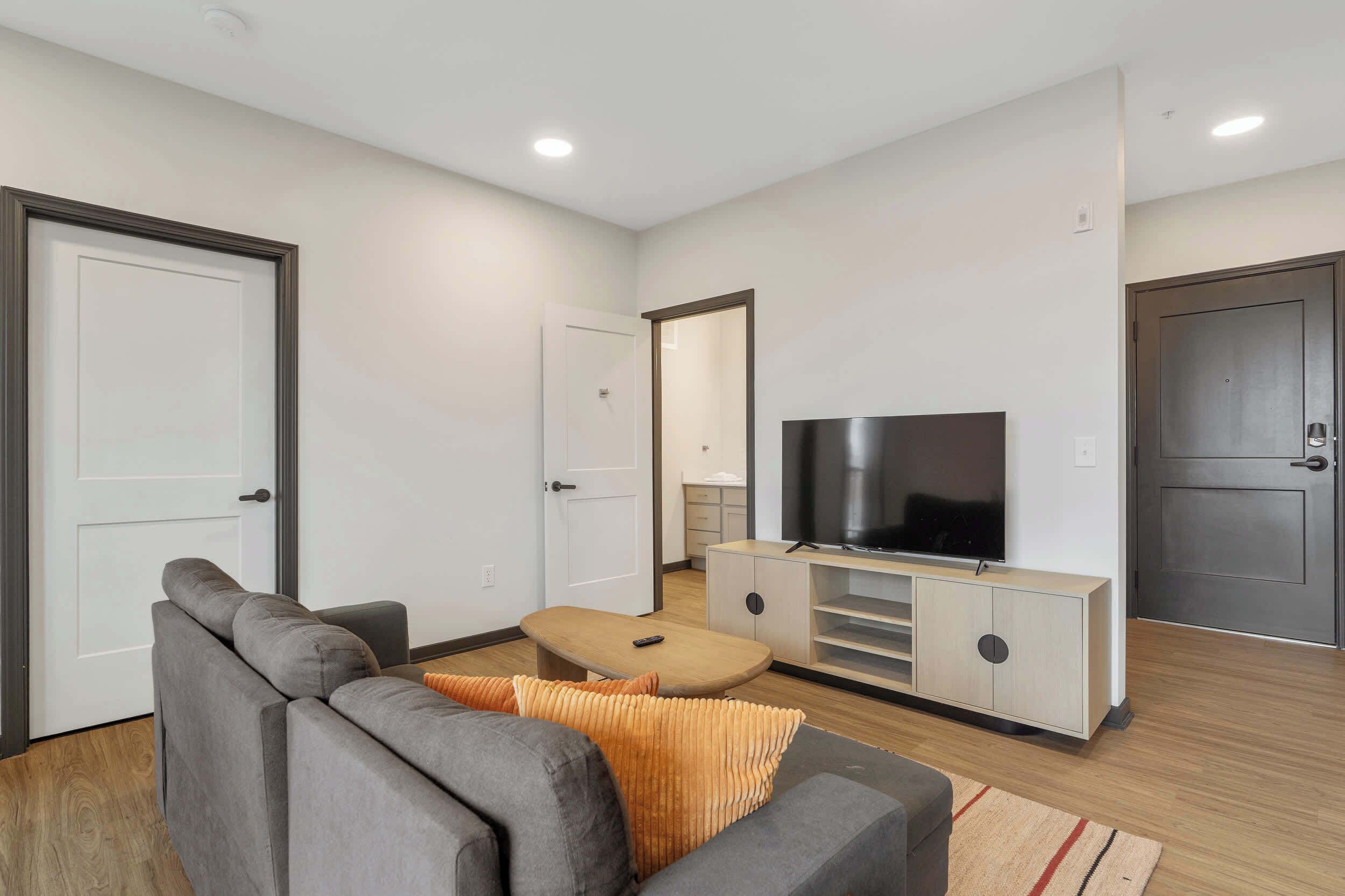 A modern living room with a grey sofa, a wooden coffee table, a TV on a wooden cabinet, and a door in the background.