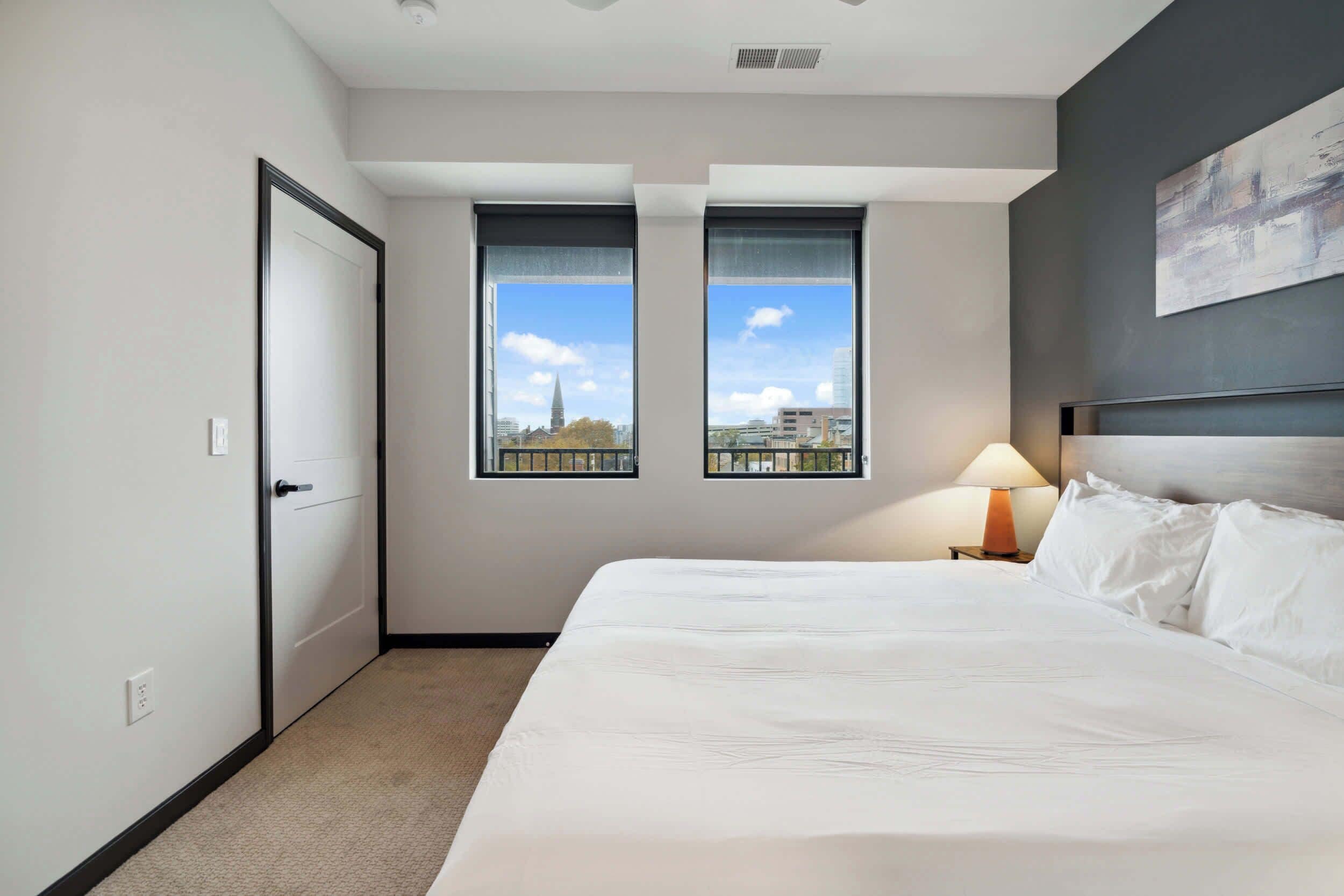 Une chambre d’hôtel avec un grand lit, deux fenêtres donnant sur la ville, une porte et une table de chevet avec une lampe.