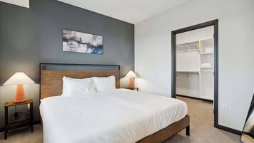 Un lit bien fait, avec du linge de lit blanc, deux lampes de chevet, une tête de lit en bois, un tableau au mur et un dressing.