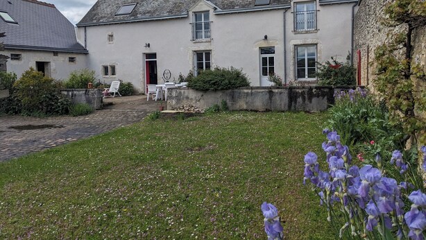 A two-story house with a paved courtyard, a red door, and a small garden with purple flowers.