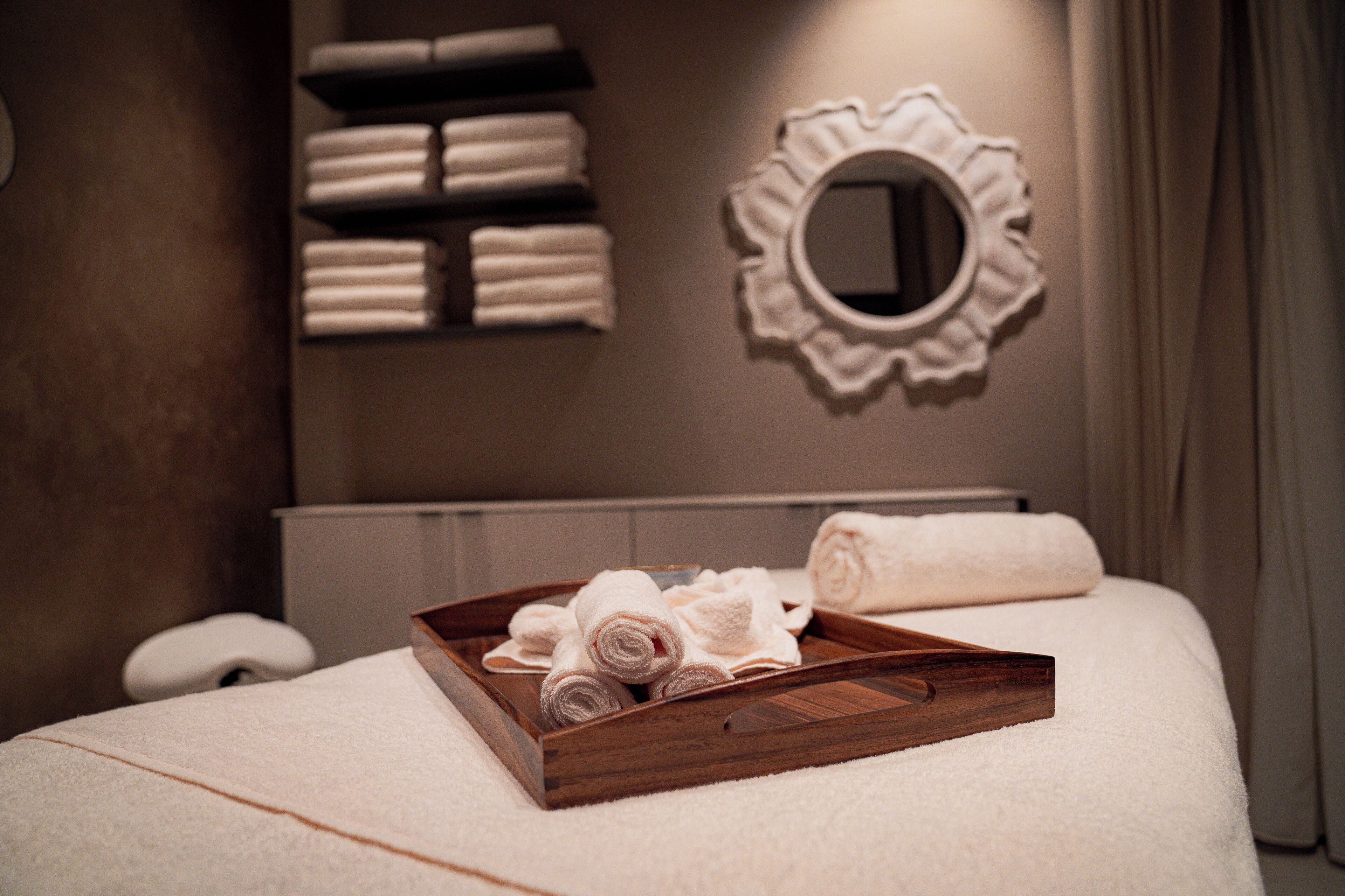 A neatly made bed with a wooden tray of rolled towels, a wall-mounted mirror, and a shelf with folded towels.