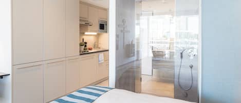 A modern hotel room with a kitchenette, a shower area, and a bed with a striped pillow.