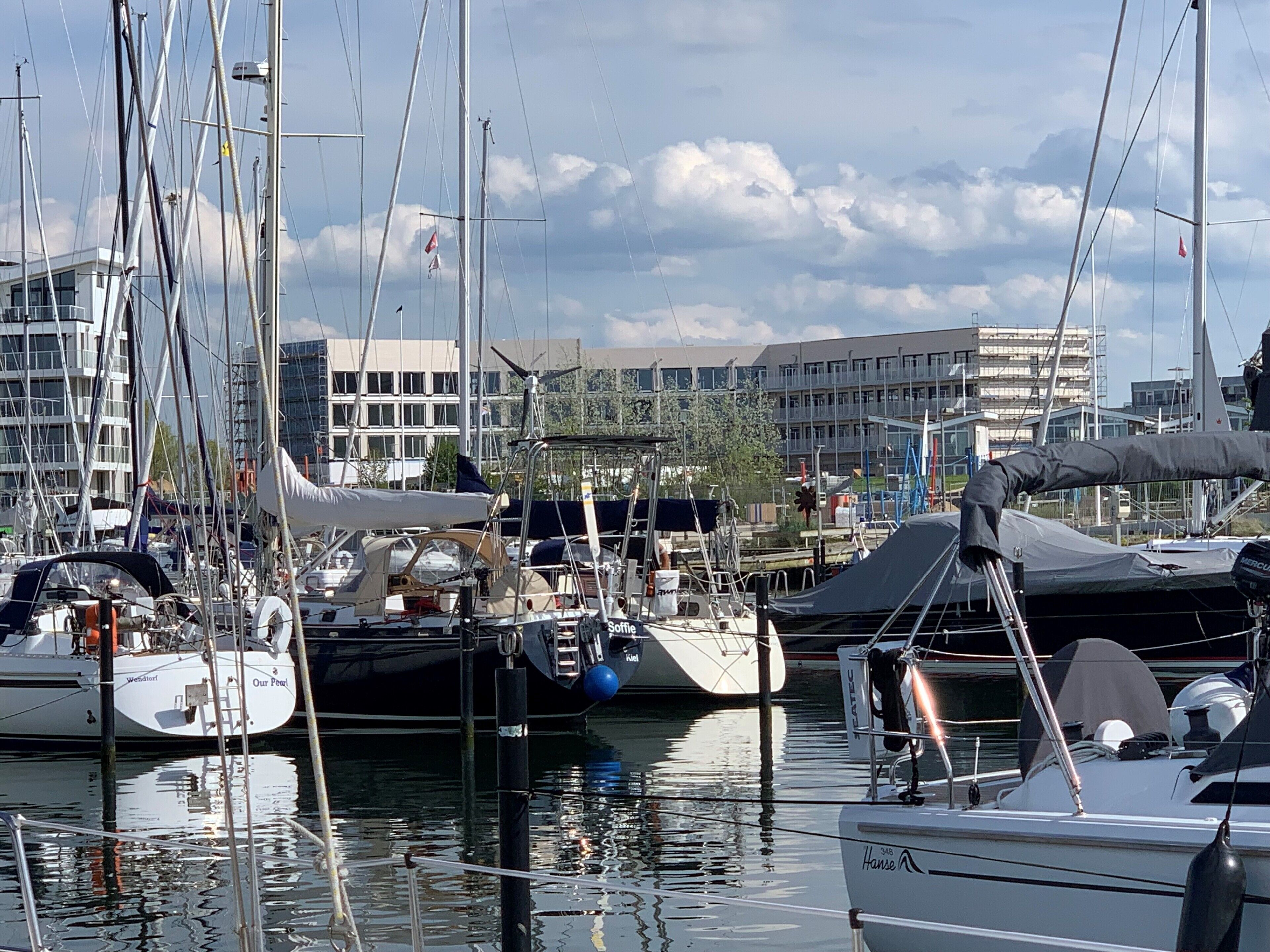 A marina with multiple sailboats docked, surrounded by buildings and trees.