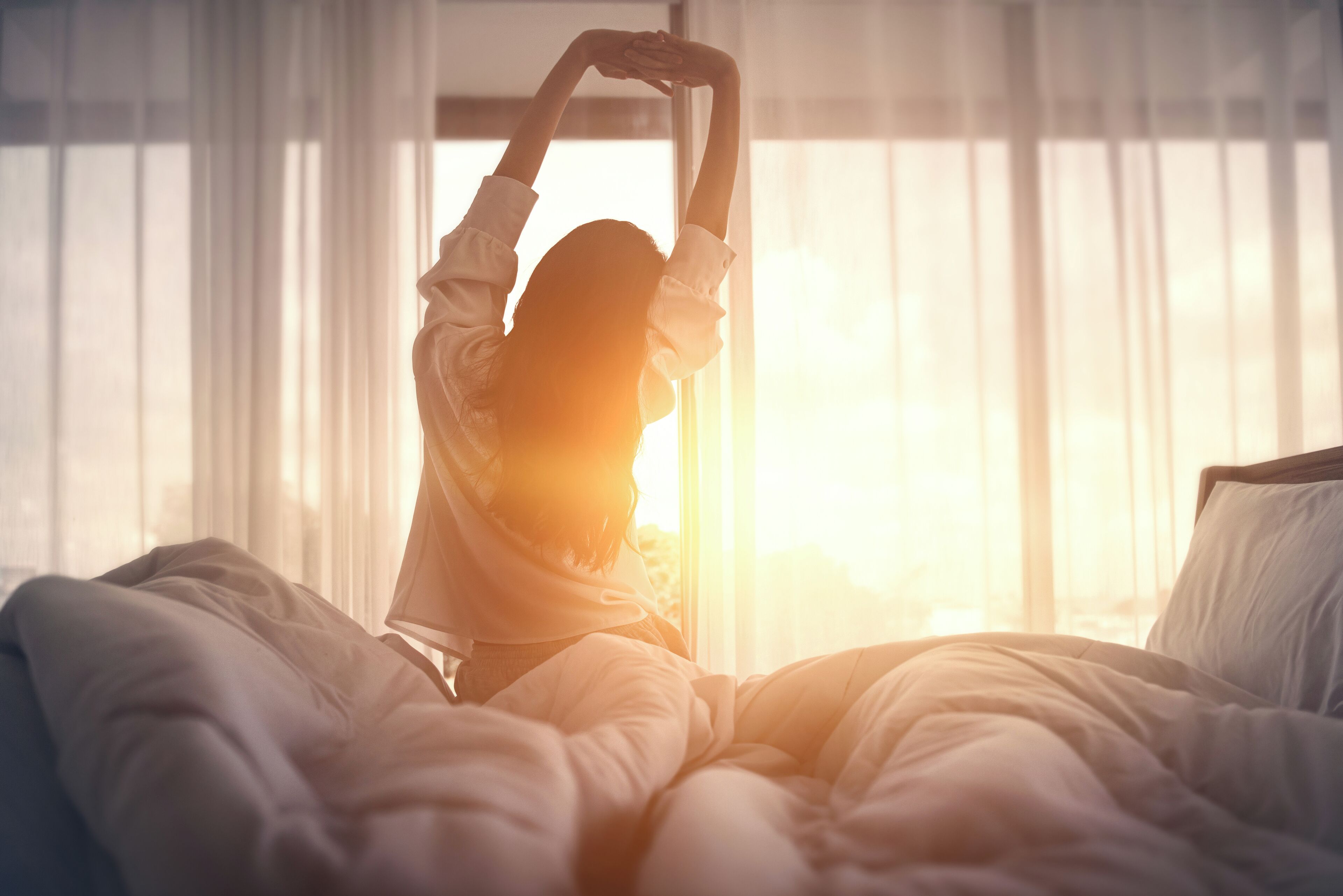 A person stretching in a room with a large window and a bed with white bedding.
