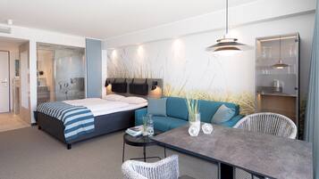 A modern hotel room with a bed, sofa, dining table, and a glass shower enclosure.