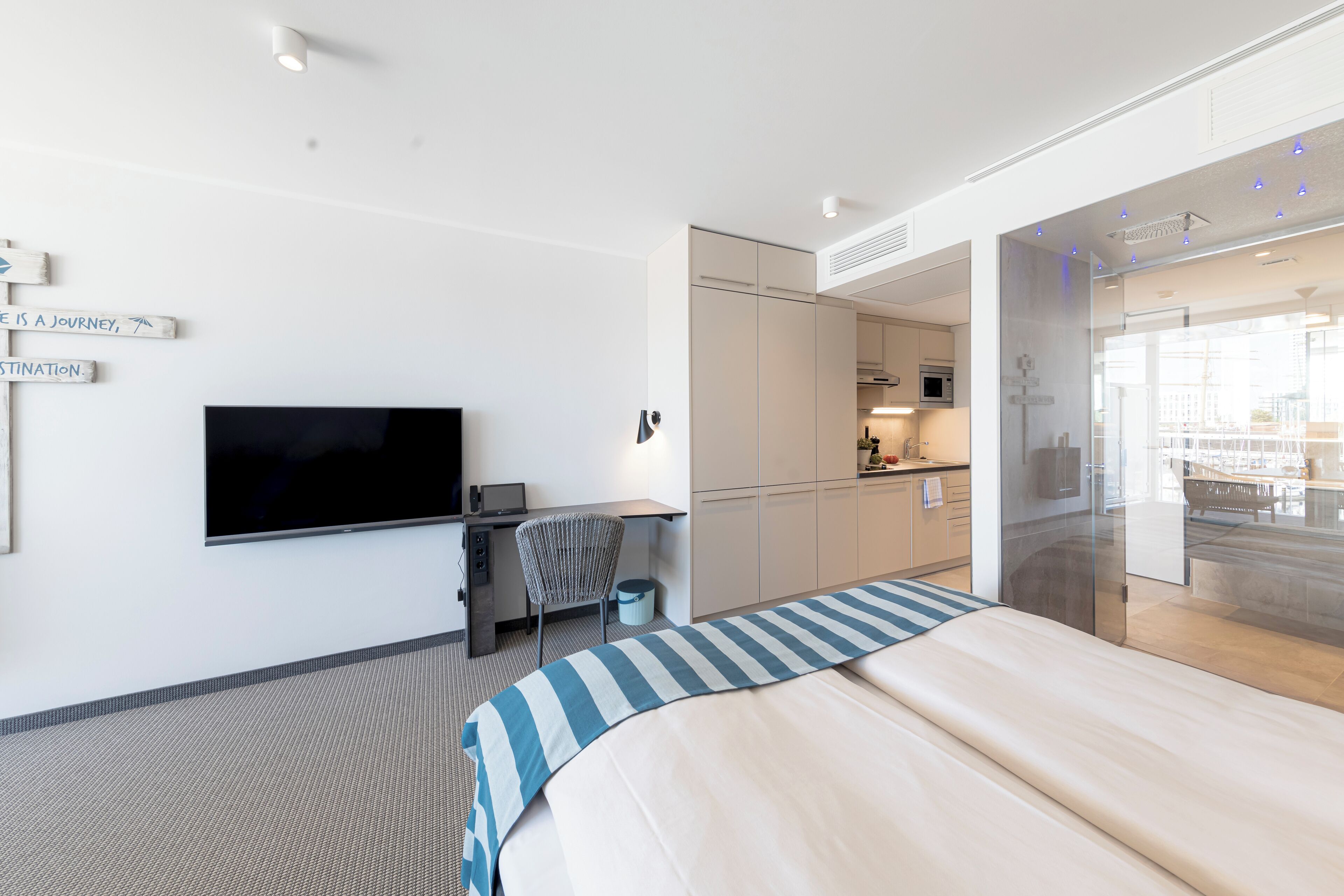 A modern hotel room with a large bed, a flat-screen TV, a desk with a chair, a kitchenette, and a glass shower area.