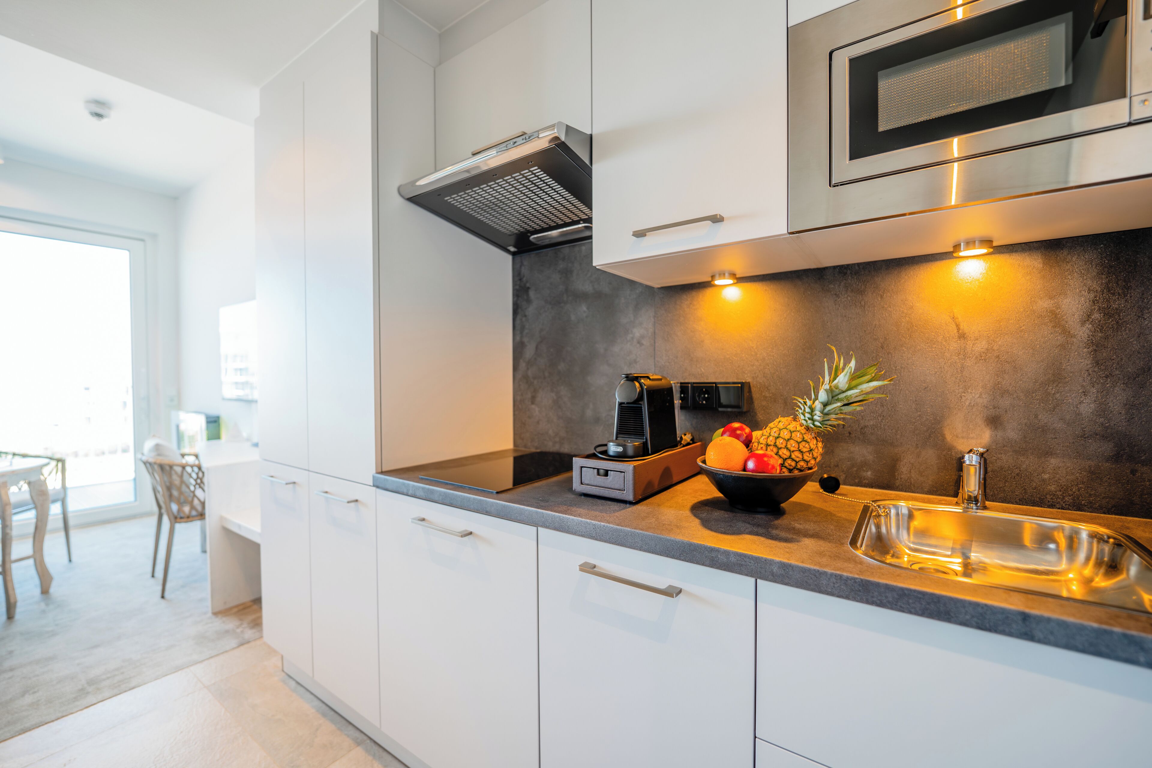 Une cuisine moderne avec des meubles blancs, un four à micro-ondes en acier inoxydable et un panier de fruits posé sur le plan de travail.