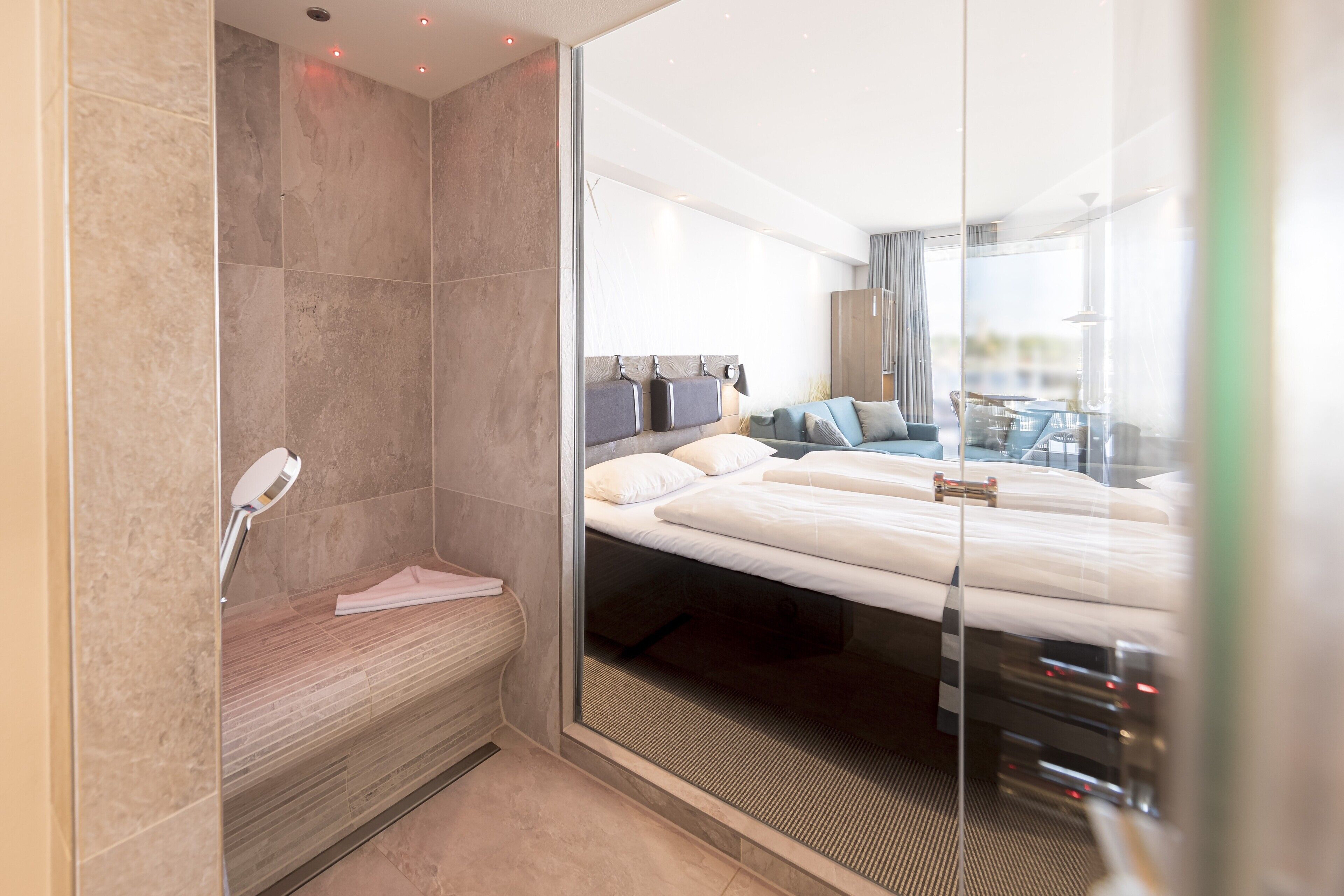 Ein modernes Hotelzimmer mit einem großen Bett, einer Sitzecke mit einer Bank und einer Glaskabine.