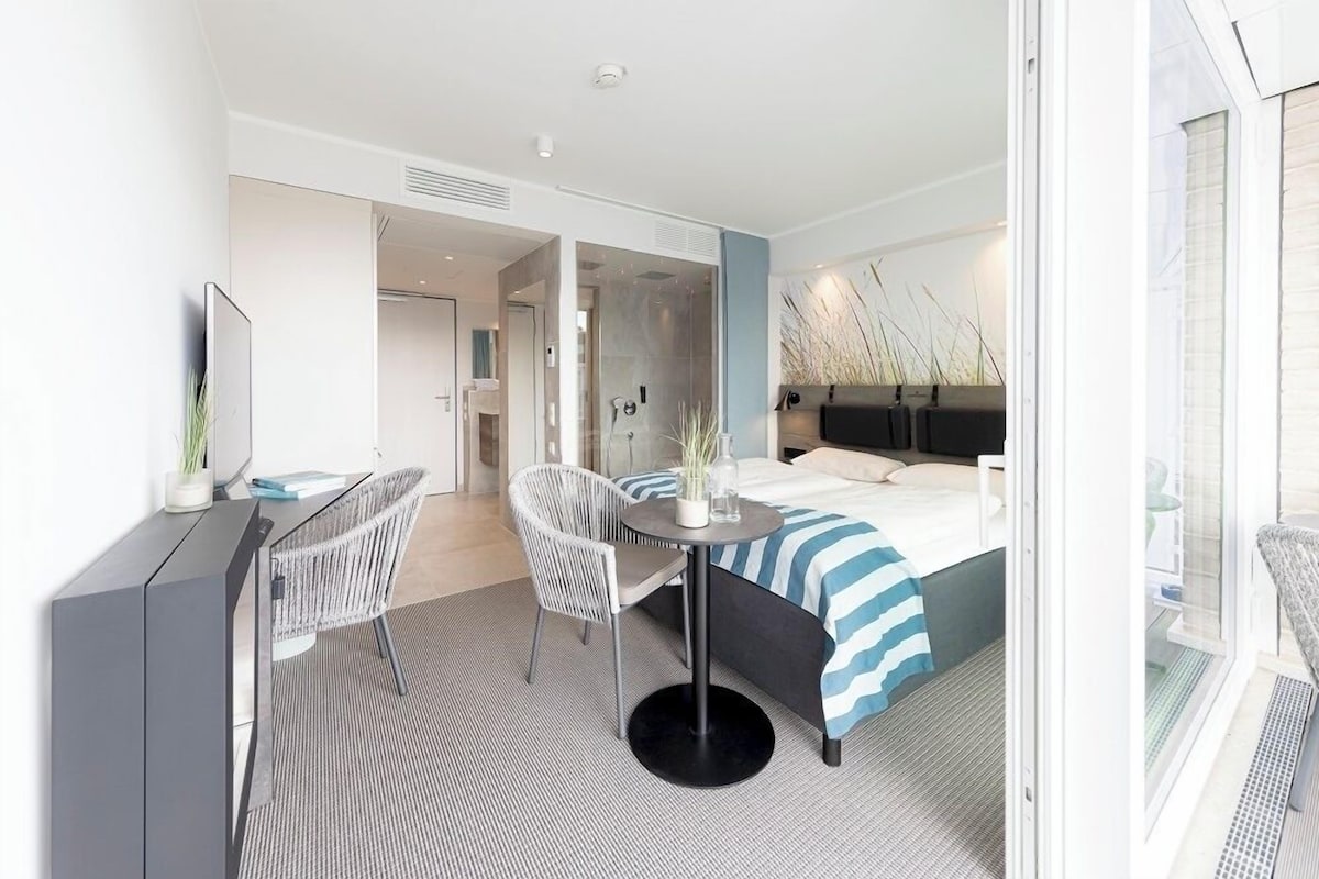 A modern hotel room with a bed, a desk, two chairs, a small table, and a large window.