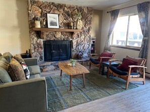 Un salon avec une cheminée en pierre, un canapé, une table basse et une vue sur l’extérieur par une fenêtre.
