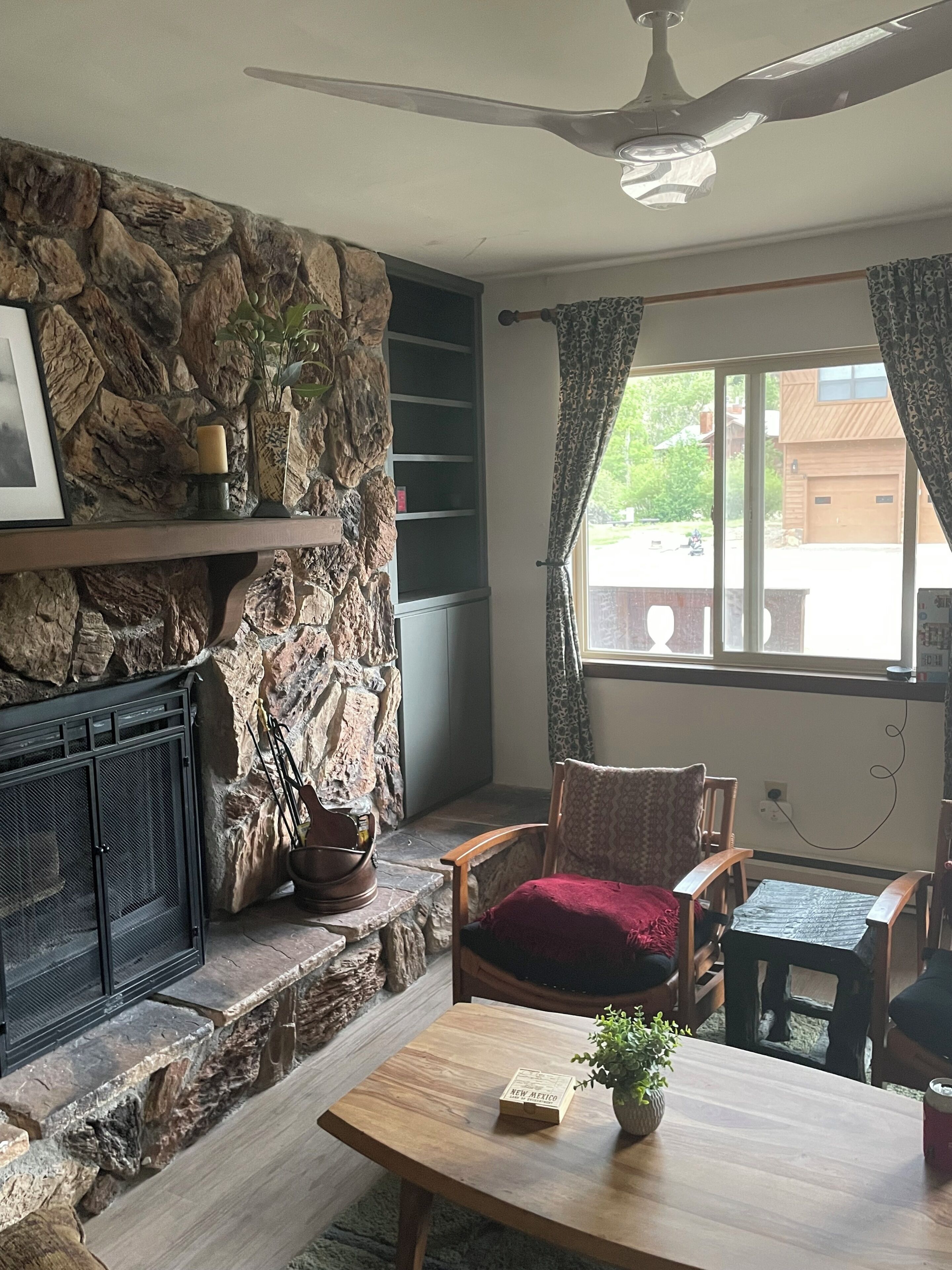 A stone fireplace, a ceiling fan, a wooden chair, a table with a book, and a window with curtains.