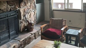 A stone fireplace, a ceiling fan, a wooden chair, a table with a book, and a window with curtains.