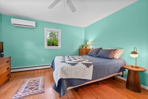 Een slaapkamer met blauwgroene muren, een bed met een bedsprei met patroon, een houten dressoir, een nachtkastje met een lamp en een raam met uitzicht op groen.