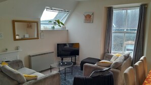 Ein Wohnzimmer mit einer Couch, Sesseln, einem Couchtisch, einem Fernseher und einem Fenster mit Vorhängen.