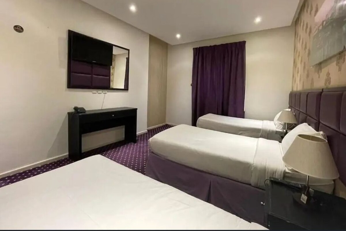Quarto de hotel com duas camas, uma escrivaninha, um espelho, um abajur e uma janela com cortinas roxas.
