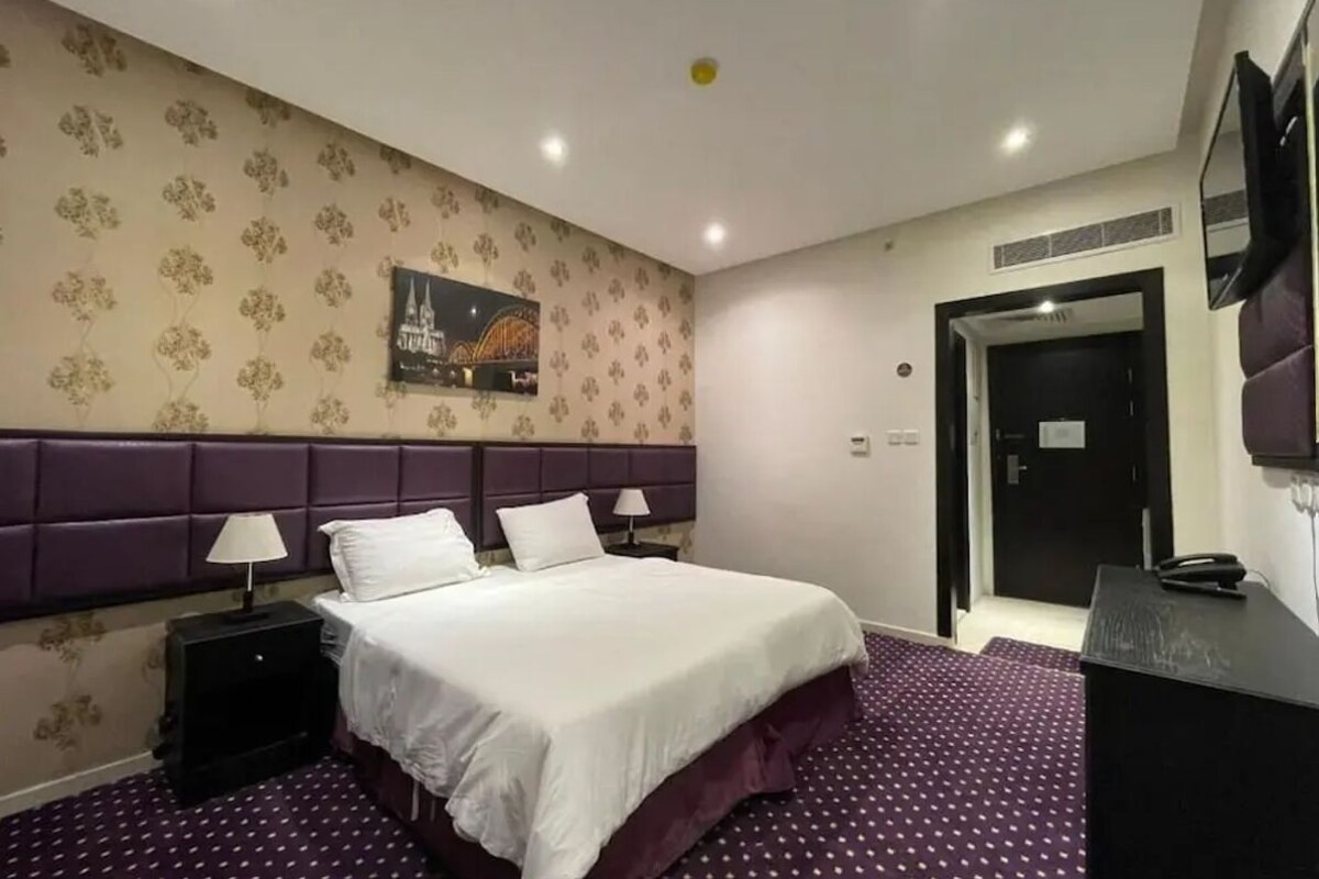 Quarto de hotel com uma cama, duas mesas de cabeceira com luminárias, uma TV fixada na parede e uma porta.