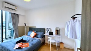 A modern bedroom with a bed, a desk, and a chair. There is a window with curtains, an air conditioning unit, and a door with a handle.