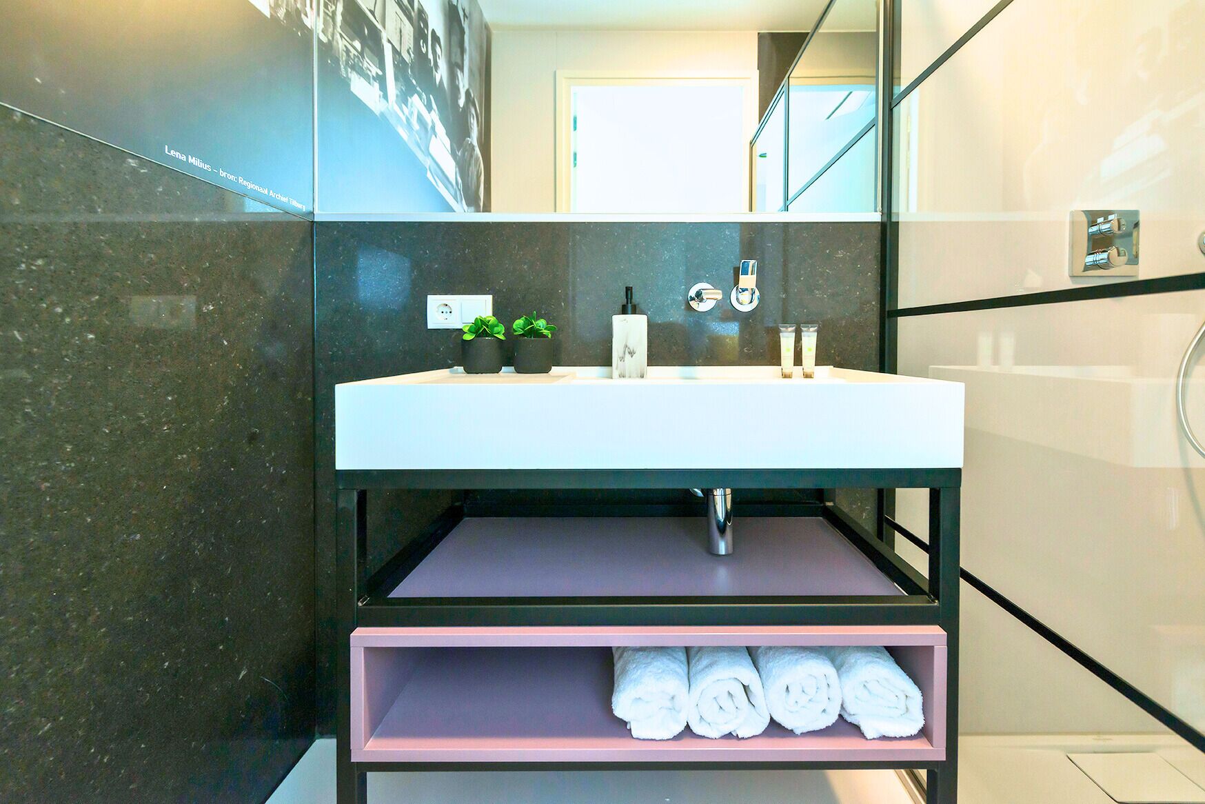 A modern bathroom with a white sink, black marble walls, a mirror, and a shelf with rolled towels.