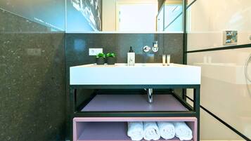 A modern bathroom with a white sink, black marble walls, a mirror, and a shelf with rolled towels.