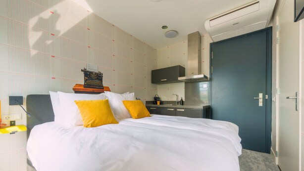A hotel room with a bed, a desk, a chair, a small kitchenette, and a door. - Unique apartment with private kitchen and bathroom, inspired by the abstract art of Lena Milius. (Tilburg)