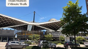 A modern train station with a distinctive roof structure, a clear sky, and a few pedestrians. - Unique apartment with private kitchen and bathroom, inspired by the abstract art of Lena Milius. (Tilburg)