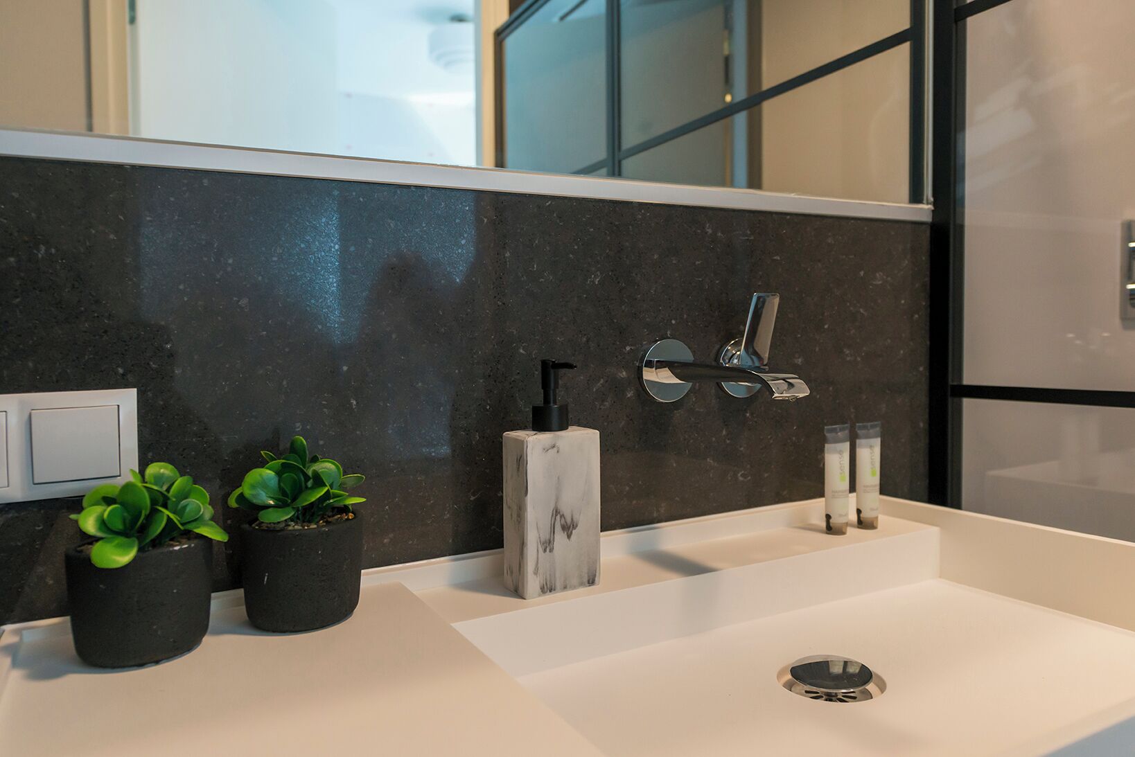 A modern bathroom with a marble backsplash, two sinks, a mirror, a faucet, and two potted plants.