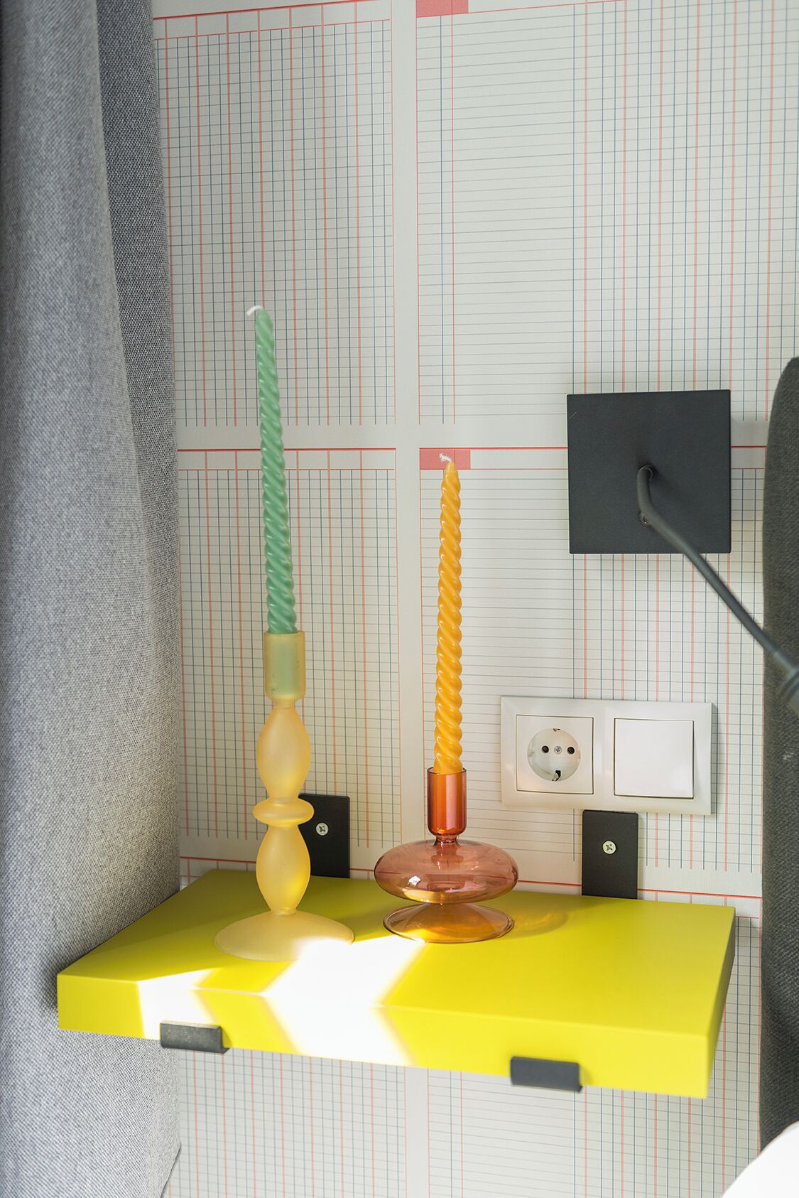 A yellow desk with two candles, a black lamp, and a power outlet on a wall with grid-patterned wallpaper.