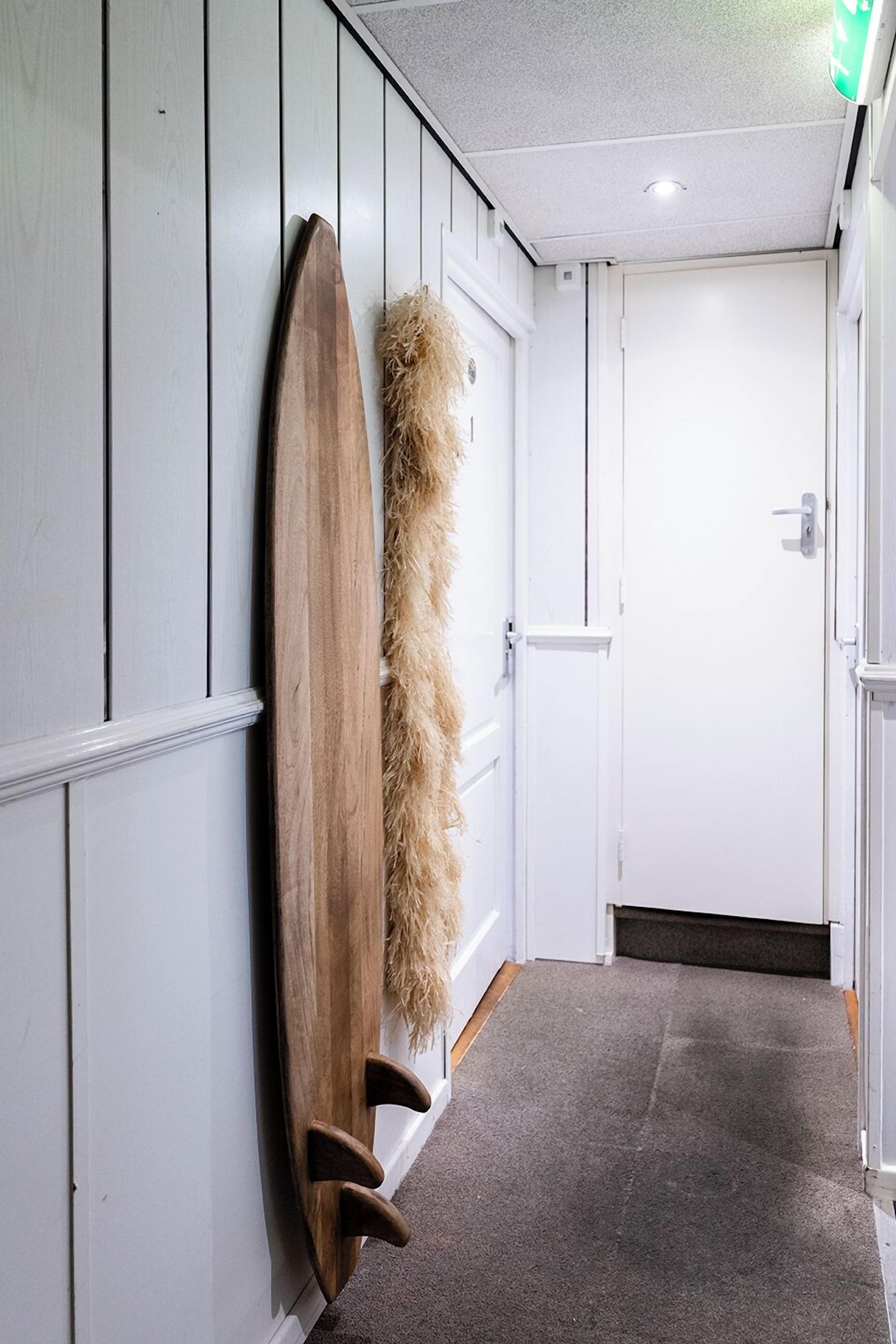 A hallway with a wooden surfboard leaning against a wall, a fluffy rug, and a closed door at the end.