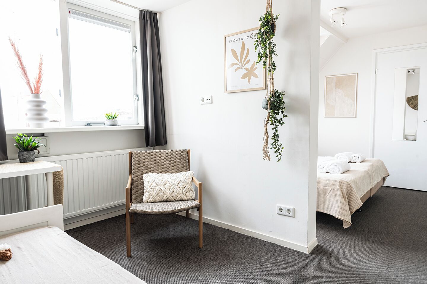 Une chambre d’hôtel moderne avec une grande fenêtre, un fauteuil en osier, un lit recouvert d’une courtepointe blanche et une plante suspendue.