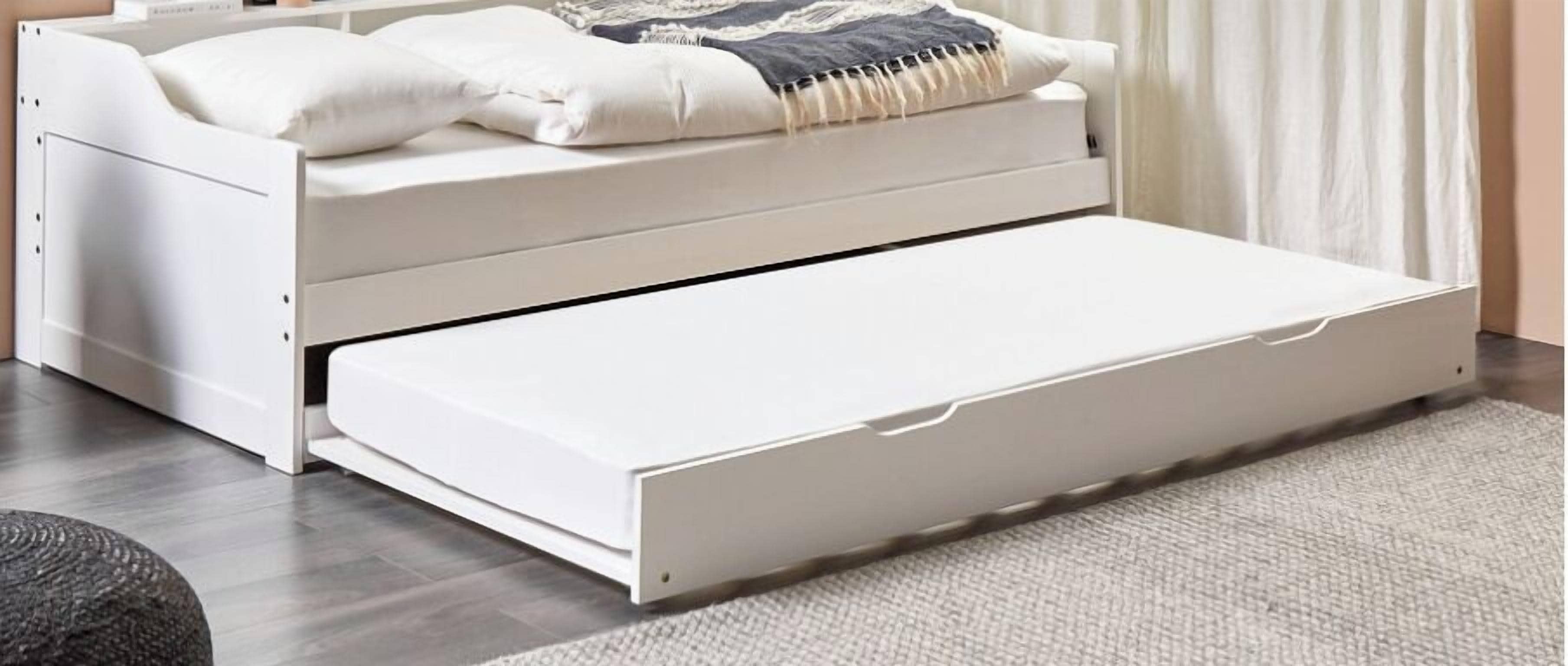 Una cama blanca con cajón extraíble, una mesita de noche y una manta decorativa.