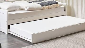 Una cama blanca con cajón extraíble, una mesita de noche y una manta decorativa.