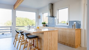 Een moderne keuken met een centraal eiland, barstoelen, grote ramen en uitzicht op groen.