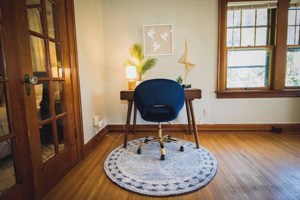 A room with a wooden floor, a round blue rug, a desk with a lamp, a chair, and a window with blinds. - Sunshine & Sky Loft in Vibrant Downtown Greeley! (Greeley)