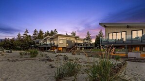 Een modern pand aan het strand met meerdere verdiepingen, grote ramen en een ruim buitengebied met zand en begroeiing.