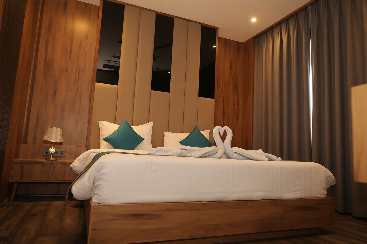 A hotel room with a large bed, wooden panel walls, a nightstand with a lamp, and a view of the outside through a window with curtains.