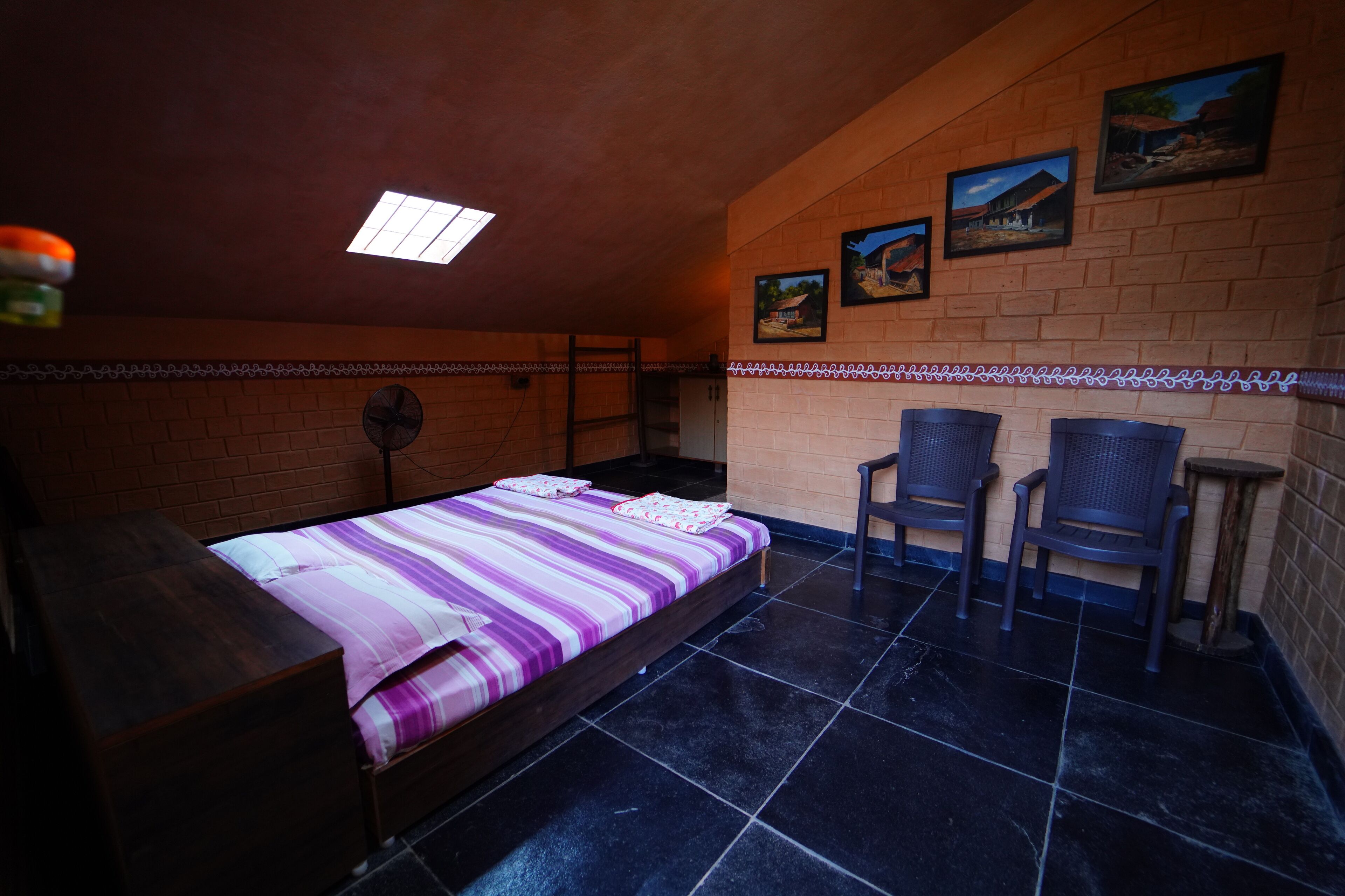 Una cama individual con colcha a rayas, una mesita, dos sillas, un ventilador y una ventana.