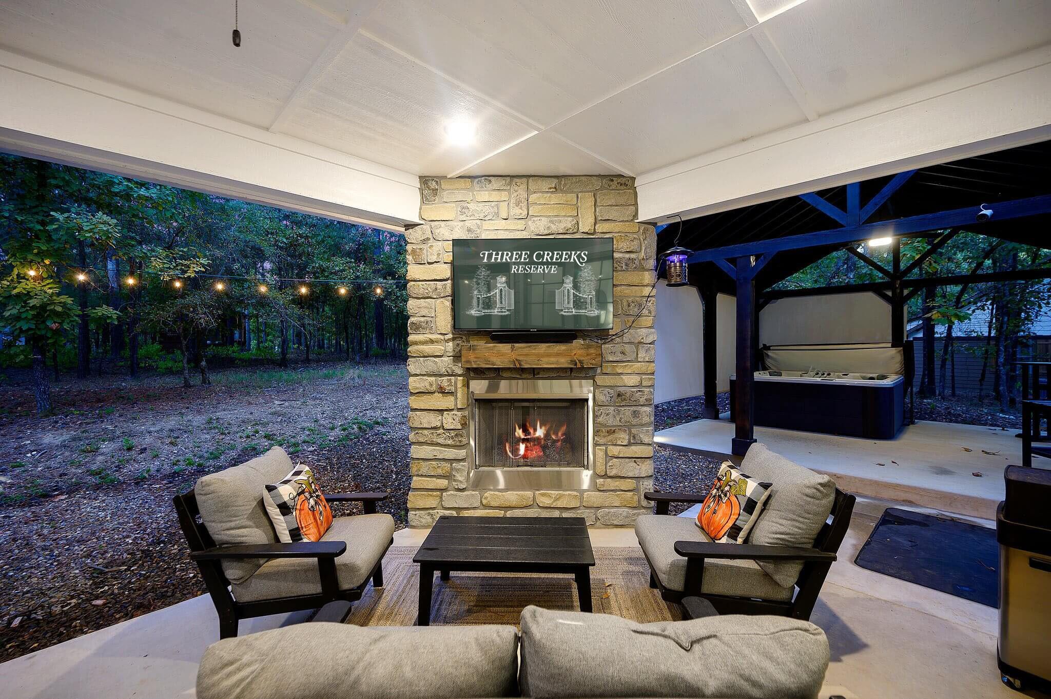 An outdoor seating area with a stone fireplace, a sign reading "Three Creeks Reserve," and a view of a forested area.