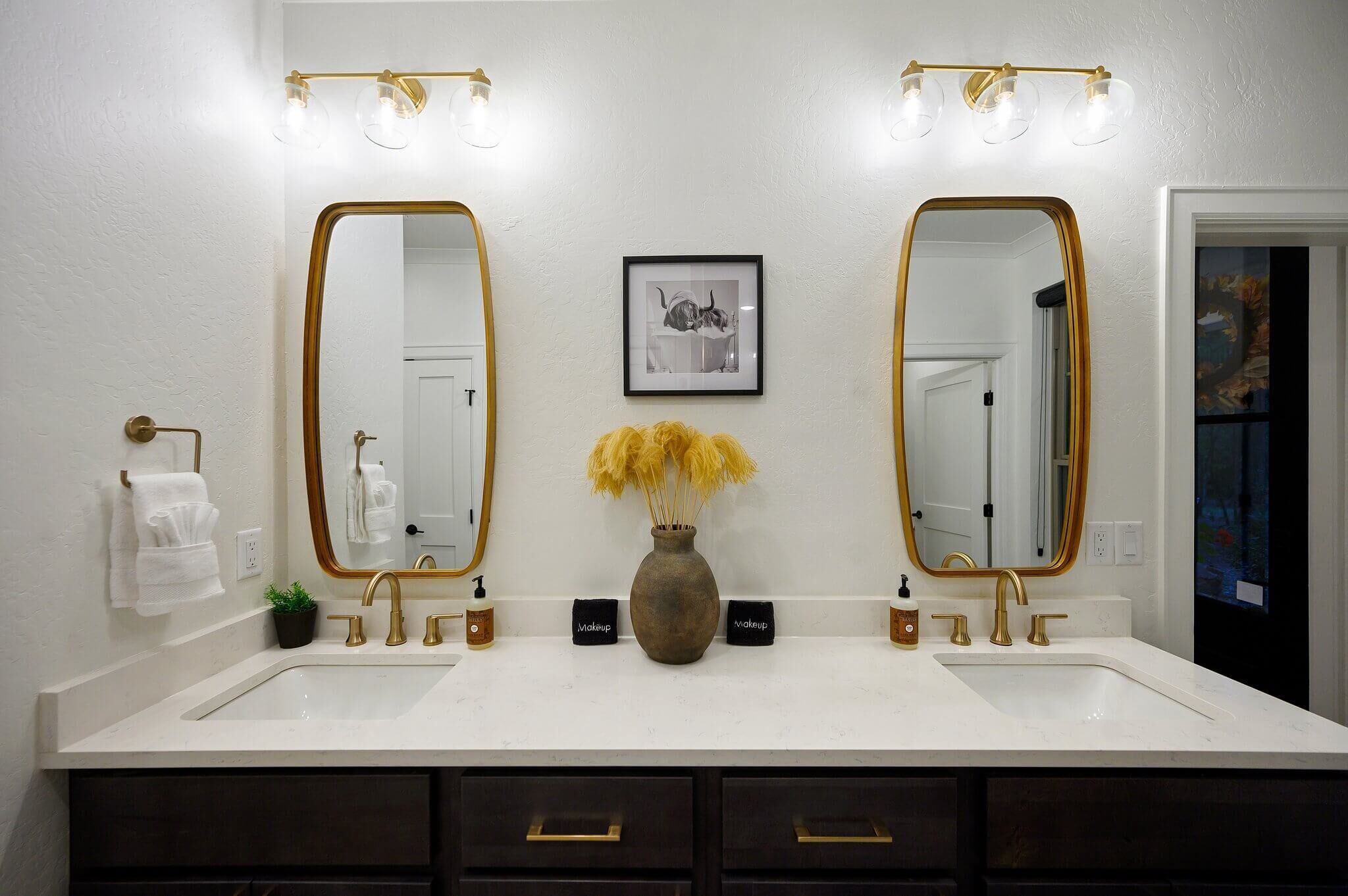 Une salle de bain avec deux lavabos, des robinets dorés et un vase contenant des fleurs séchées.