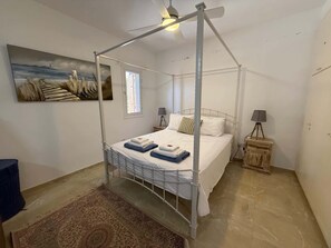 A four-poster bed with a ceiling fan, a painting on the wall, a small bedside table with a lamp, and a rug on the floor. - Regina Gardens - Spacious, 3BR, Ground Floor Apart. with Huge Patio (Paphos)
