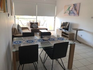 Una sala de estar moderna con una mesa de comedor de vidrio, sillas negras, un sofá, un televisor y una pequeña mesita auxiliar.
