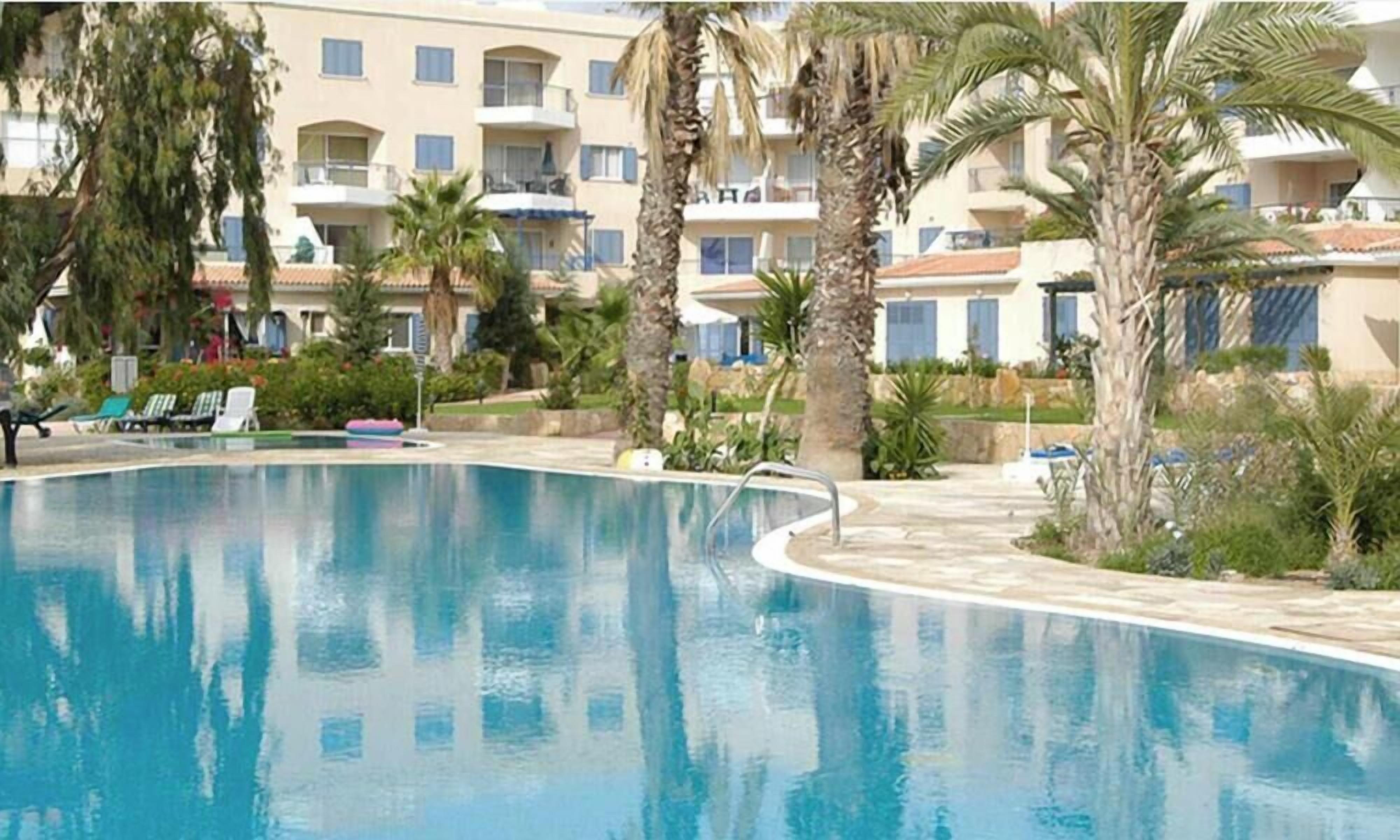 Piscina con sillas de descanso y palmeras, rodeada por un edificio con balcones.